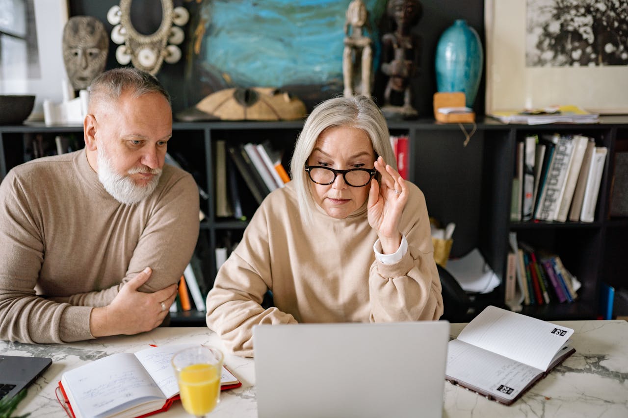 An elderly couple in a cozy home setting engaged with a laptop, planners, and a glass of juice on the table.