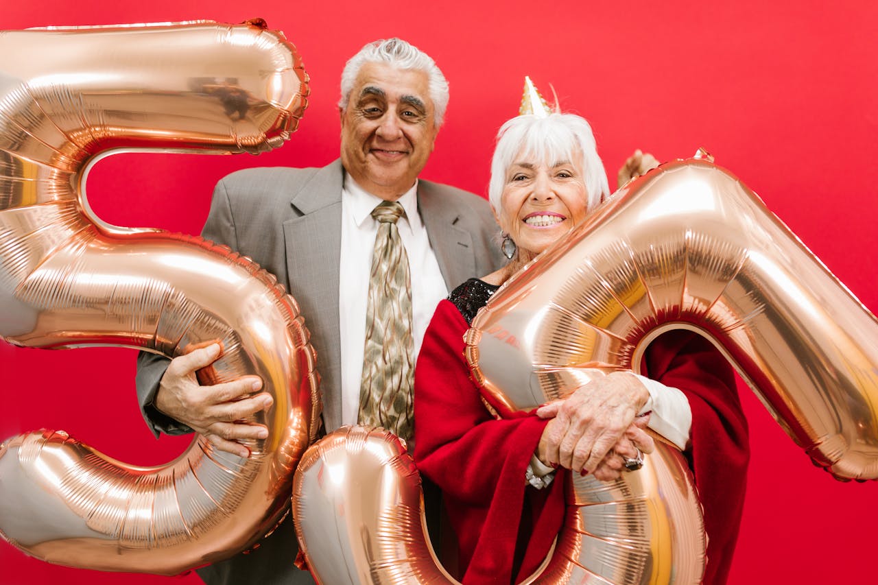 Cheerful senior couple celebrating with gold balloons on a vibrant red background.